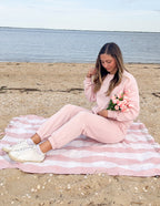 Woman seated on the beach wearing a pink Christian Crewneck Sweatshirt, holding flowers by the water
