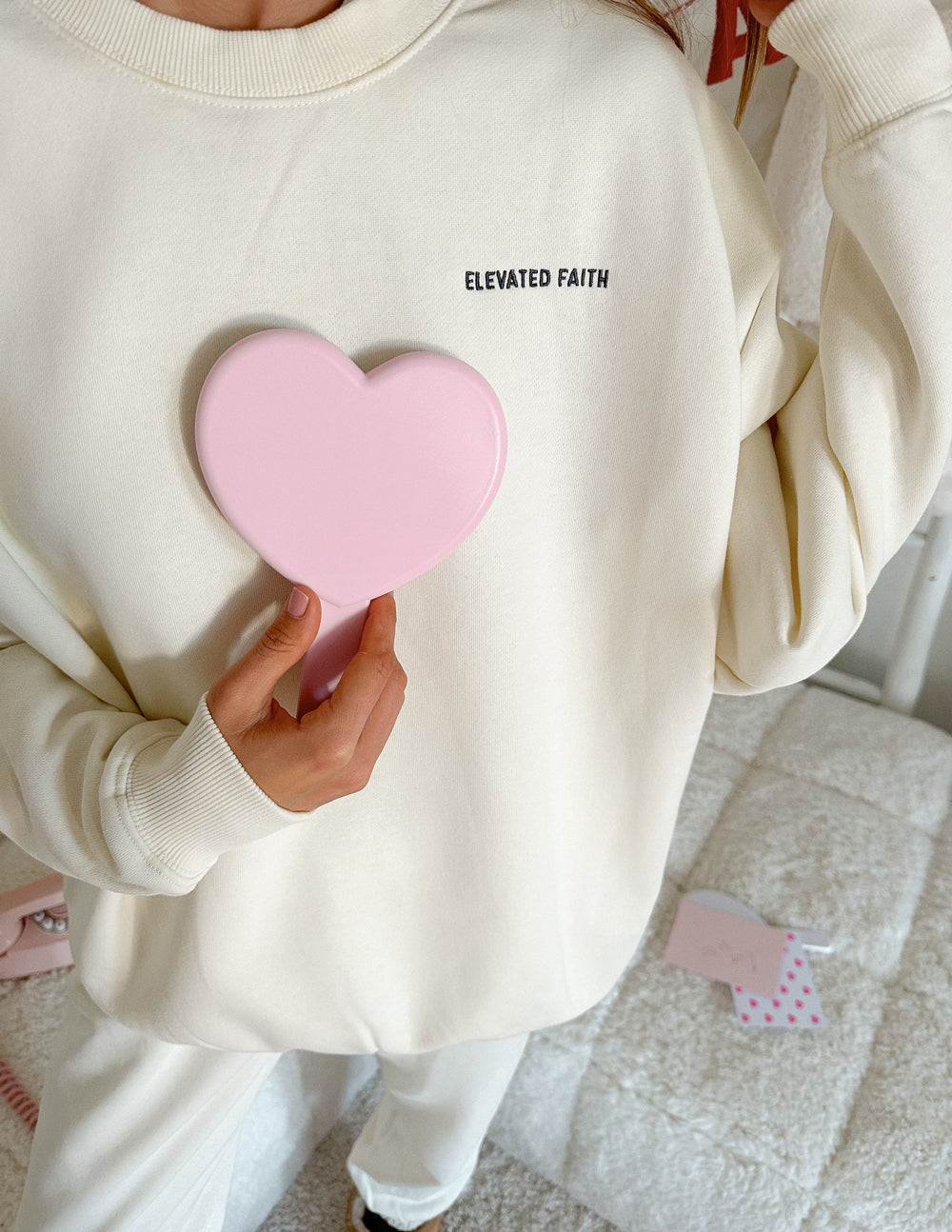 Model wearing an Elevated Faith Christian Crewneck Sweatshirt in cream color, holding a pink heart mirror.