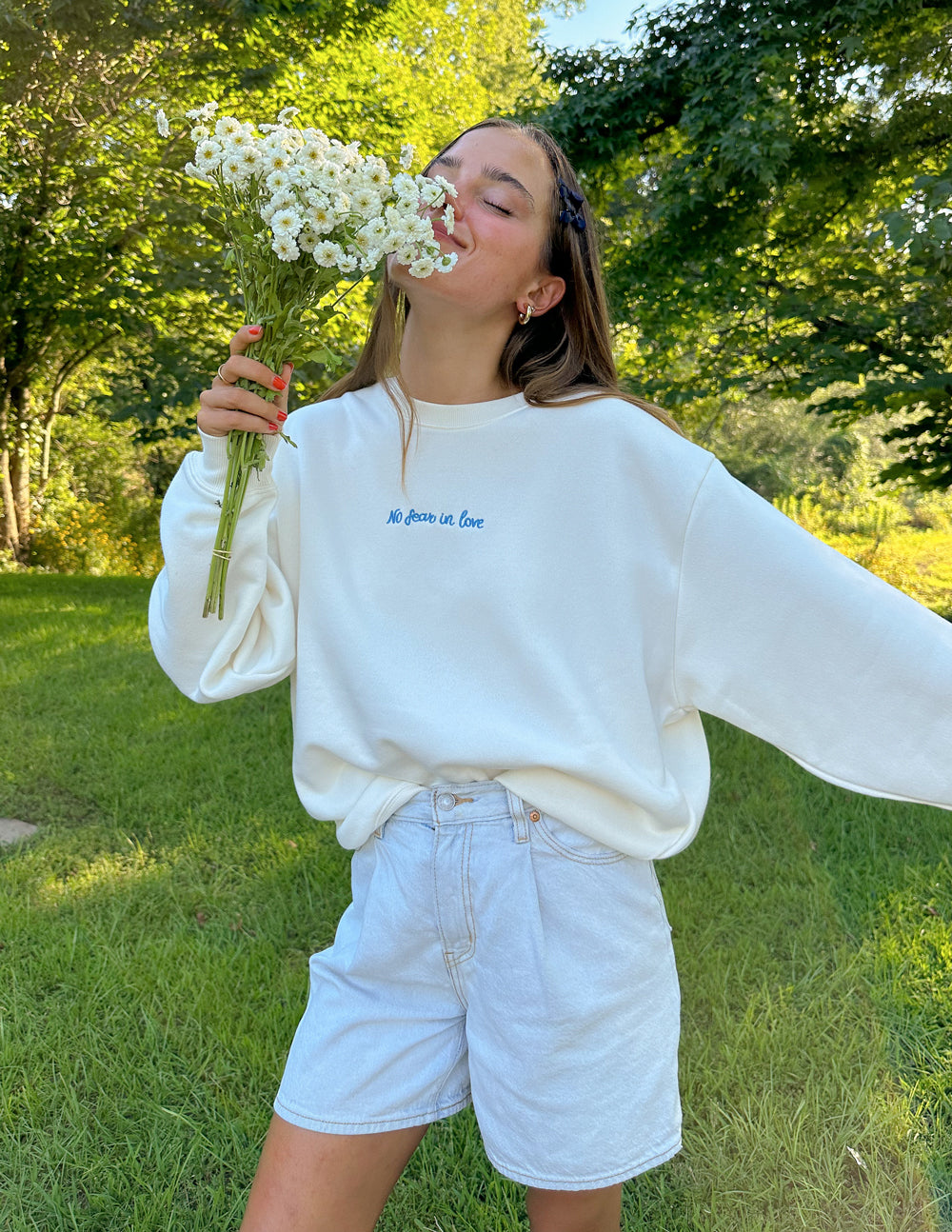 Model wearing a white Christian Crewneck Sweatshirt with floral details in a green outdoor setting