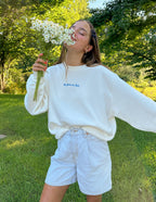 Model wearing a white Christian Crewneck Sweatshirt with floral details in a green outdoor setting