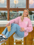 Woman wearing a pink Christian Crewneck Sweatshirt sitting with iced coffee at a cafe