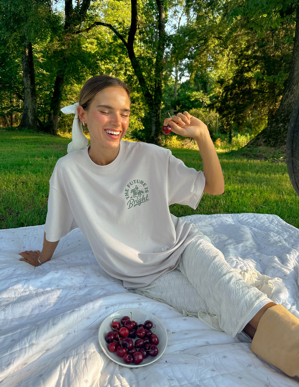 Elevated Faith Christian Apparel T-Shirt in light color, worn by a smiling woman outdoors, sitting on a blanket