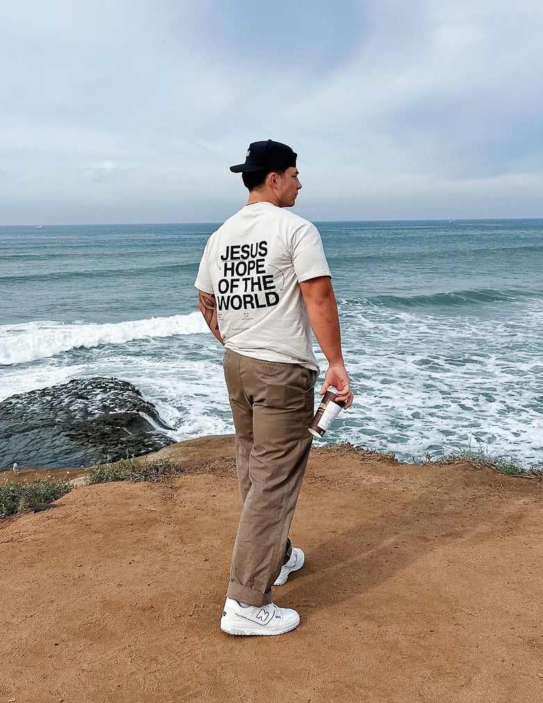 Man wearing Elevated Faith Christian Apparel T-Shirt with 'Jesus Hope of the World' on the back by the ocean
