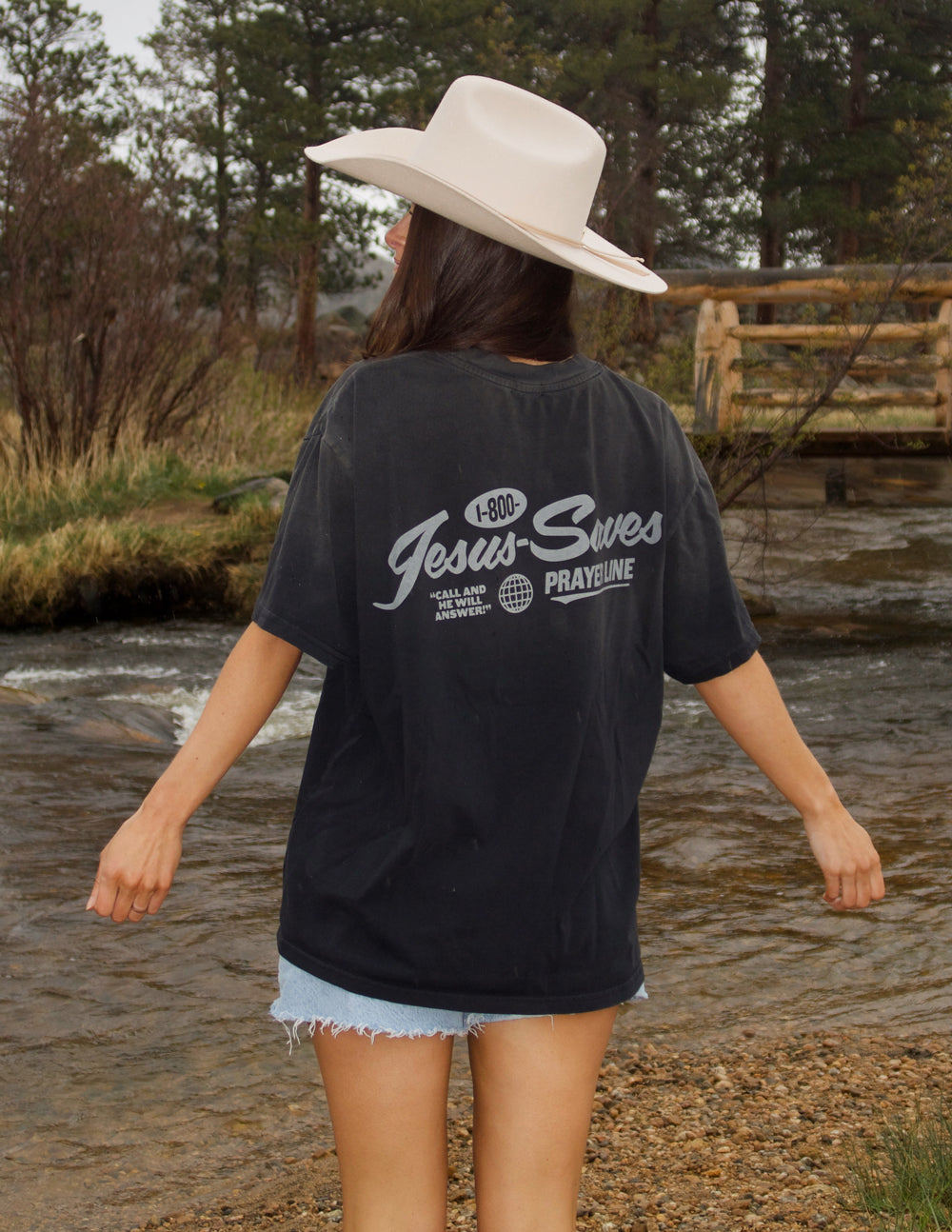 Woman wearing a black Christian Apparel T-Shirt with 'Jesus Saves' design near a creek