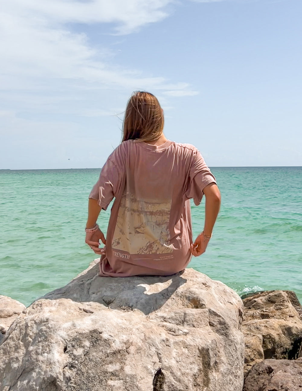 Woman wearing Elevated Faith Christian Apparel T-Shirt on rocks by the sea with a scenic backdrop