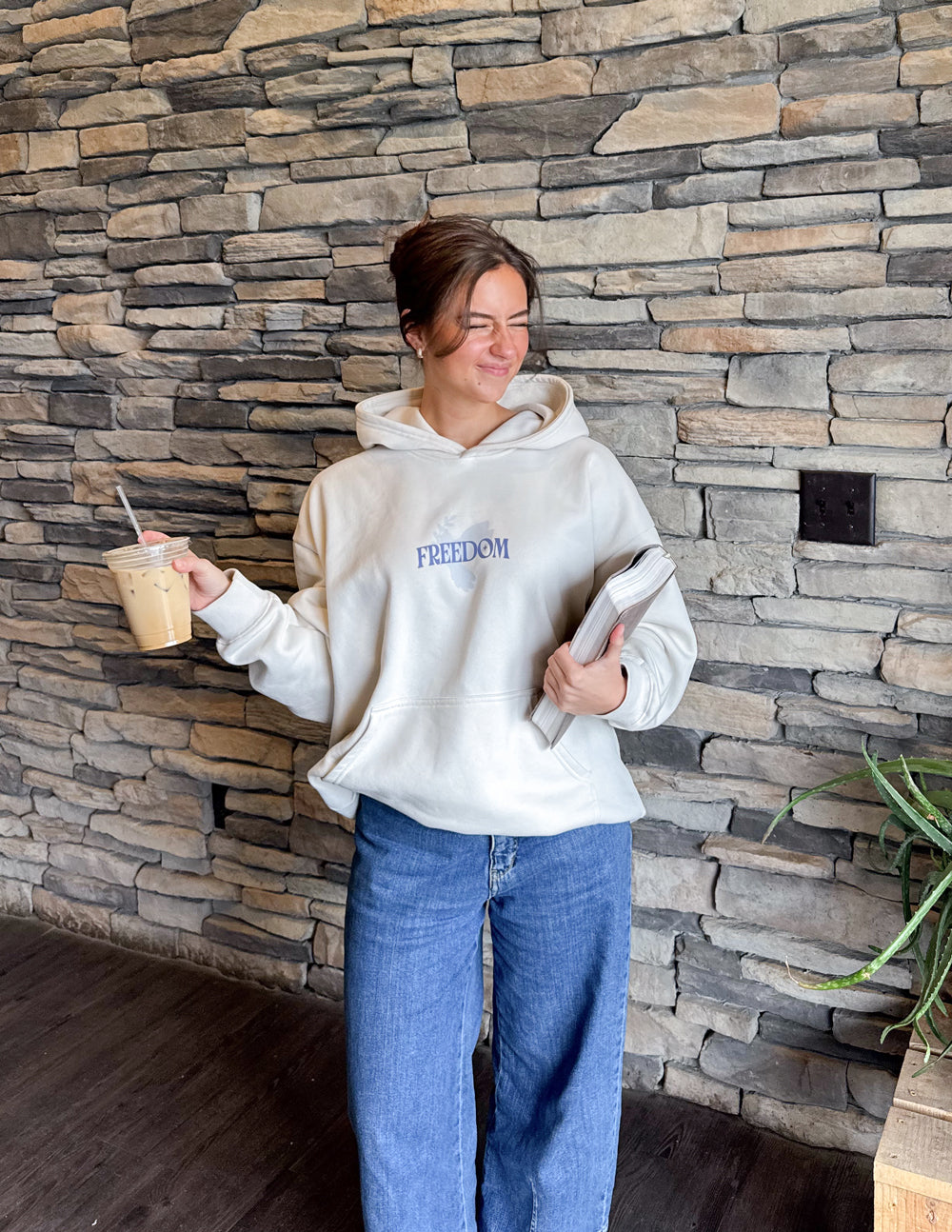 Woman wearing a beige Christian Hoodie with 'FREEDOM' print, holding coffee and a magazine