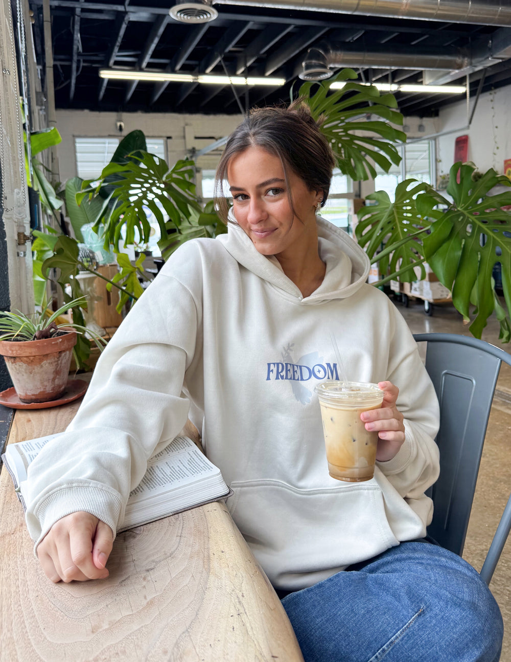 Woman in a cream Christian Hoodie with Freedom print, enjoying coffee in a cozy café