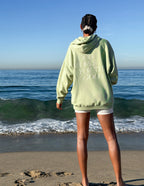 Light green Christian Hoodie by Elevated Faith with 'God works for the good' text on back at the beach