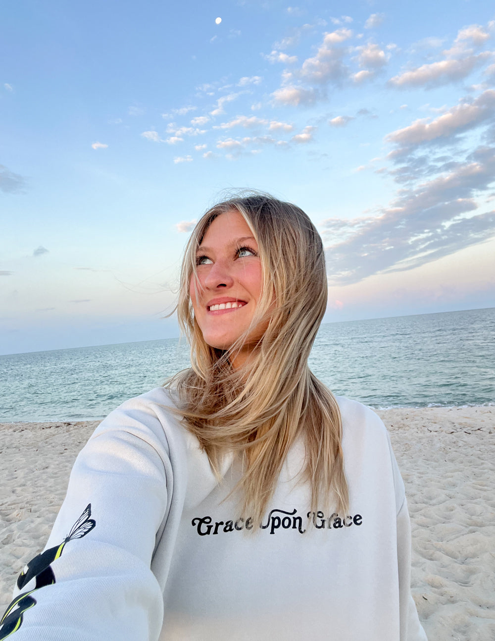 Woman wearing a Christian Hoodie with the text Grace upon Grace, standing on the beach at sunset