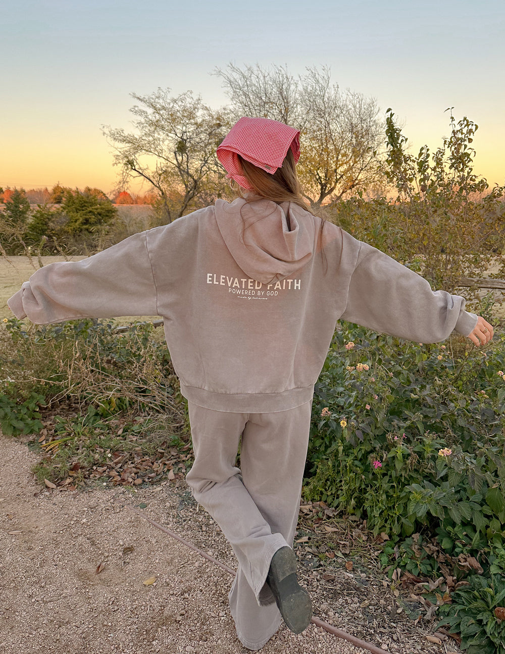 Model wearing Elevated Faith Christian Hoodie with oversized fit and stylish design against a sunset background