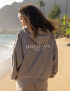 Elevated Faith Christian Hoodie in gray featuring 'Powered by God' graphic on the back by the beach