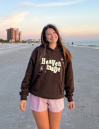 Woman wearing a brown Christian hoodie with 'Heaven made' graphic on a beach at sunset