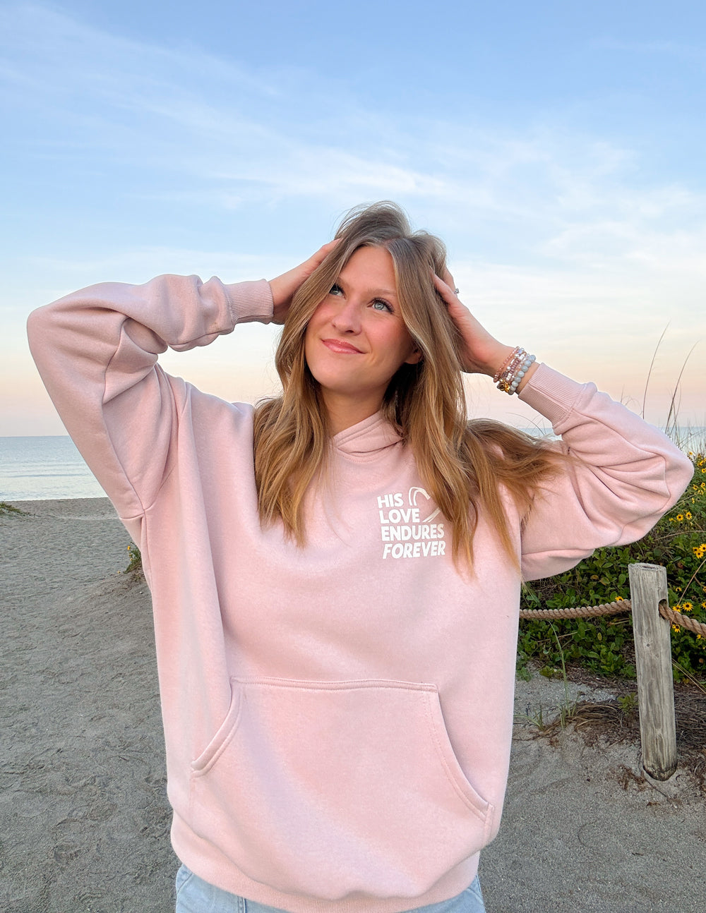 Elevated Faith Christian Hoodie in Light Pink with 'His Love Endures Forever' Design at the Beach