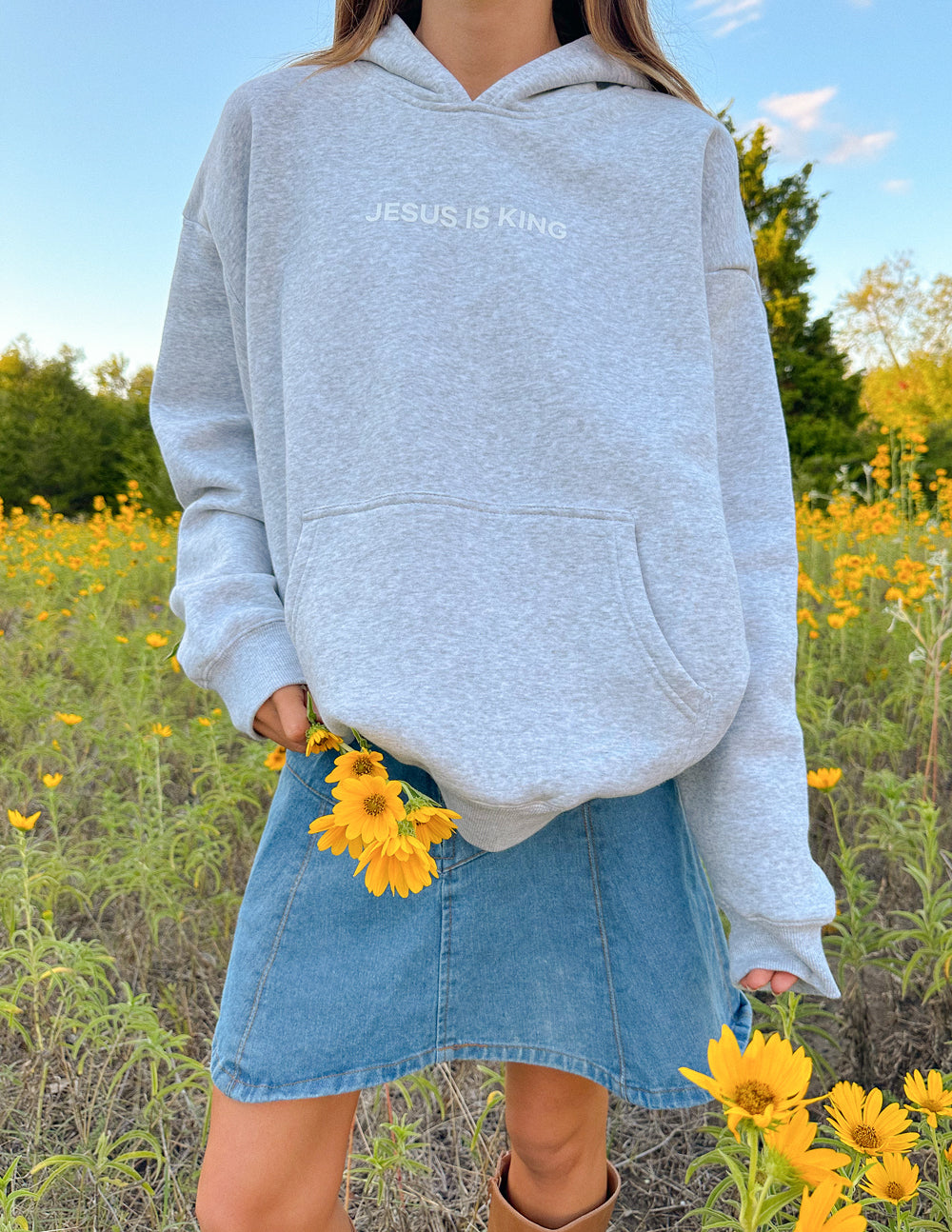 Gray Elevated Faith Christian Hoodie featuring 'Jesus is King' text, worn with a denim skirt in a sunny field