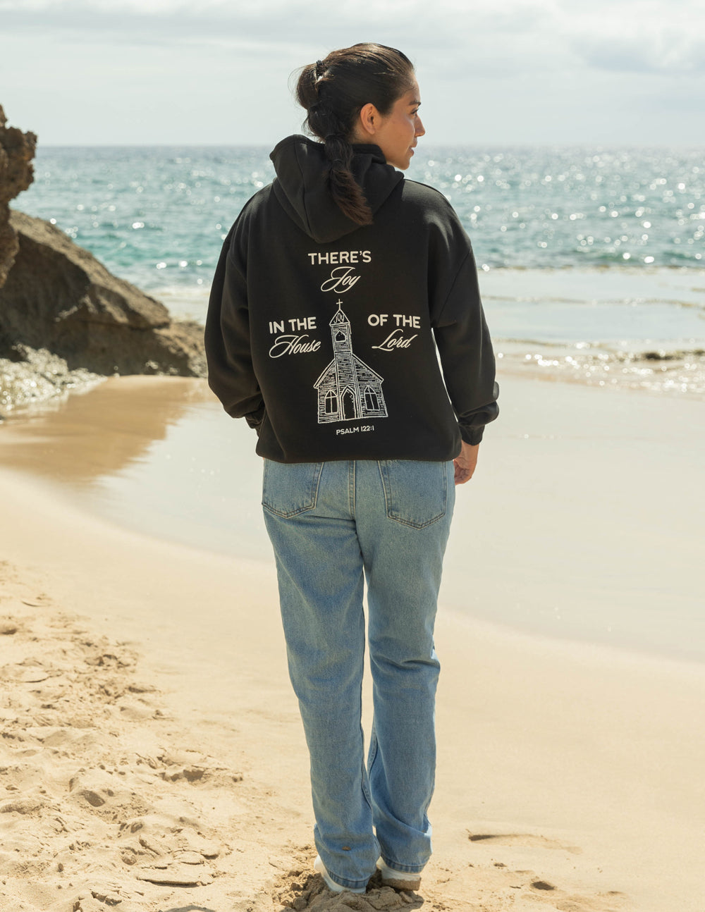 Woman wearing a black Christian Hoodie with church graphic on the back by Elevated Faith, on a beach