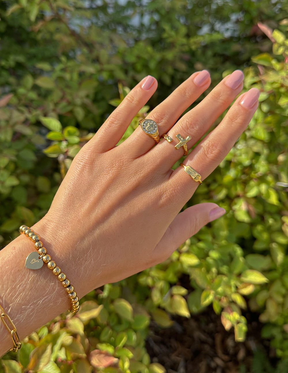 Elegant hand wearing Elevated Faith Christian Jewelry rings and a gold bracelet among greenery