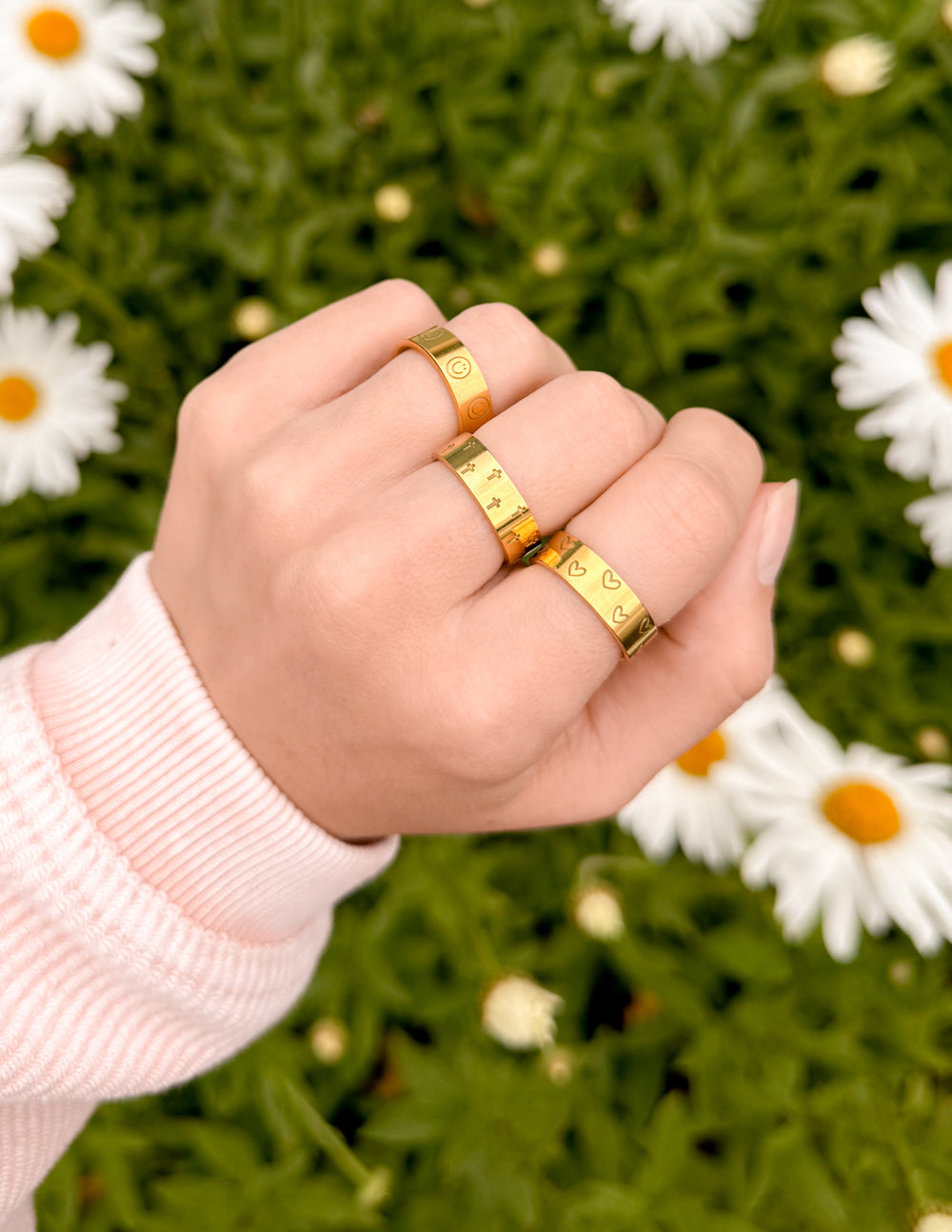 Three gold rings displaying Christian Jewelry symbols worn on a hand among daisies