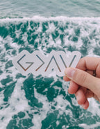Hand holding a transparent sticker with the word 'GRAV' above ocean waves, great for Christian Stickers.